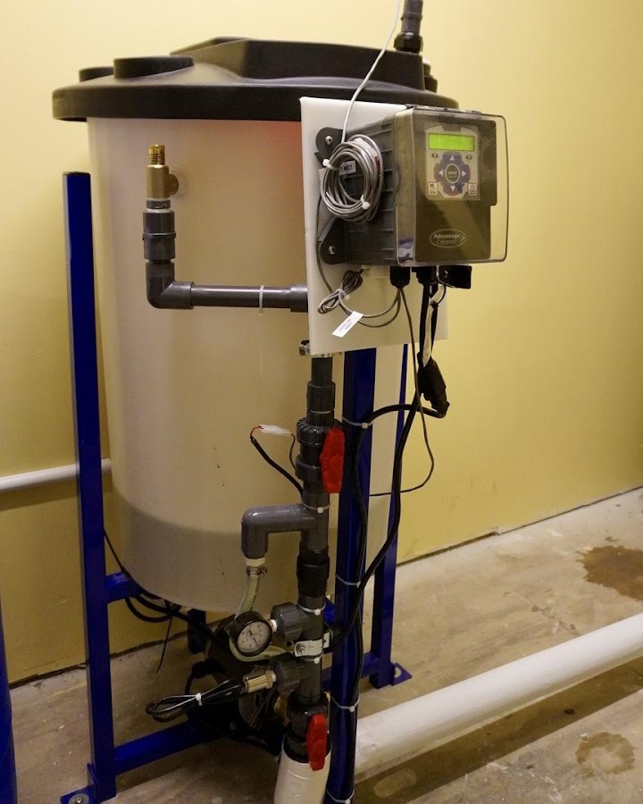 A boiler water treatment chemical feed tank with a digital controller and gauge, installed in a mechanical room.