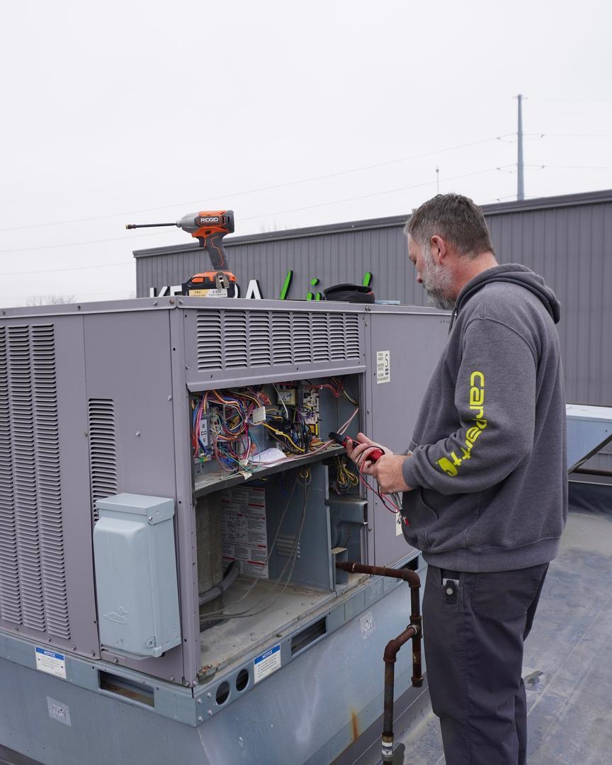 Ohio Heating technician taking electrical readings inside an open packaged rooftop HVAC unit on a flat commercial roof in Columbus